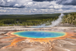 Grand Prismatic Spring in Yellowstone, with blue and turquoise water, orange edges, rising steam and dramatic clouds above the forest. Photograph by Simone Amaduzzi.